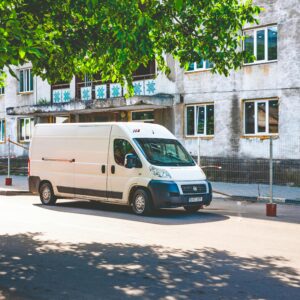 A white delivery van parked under a tree on a sunny day in an urban area.