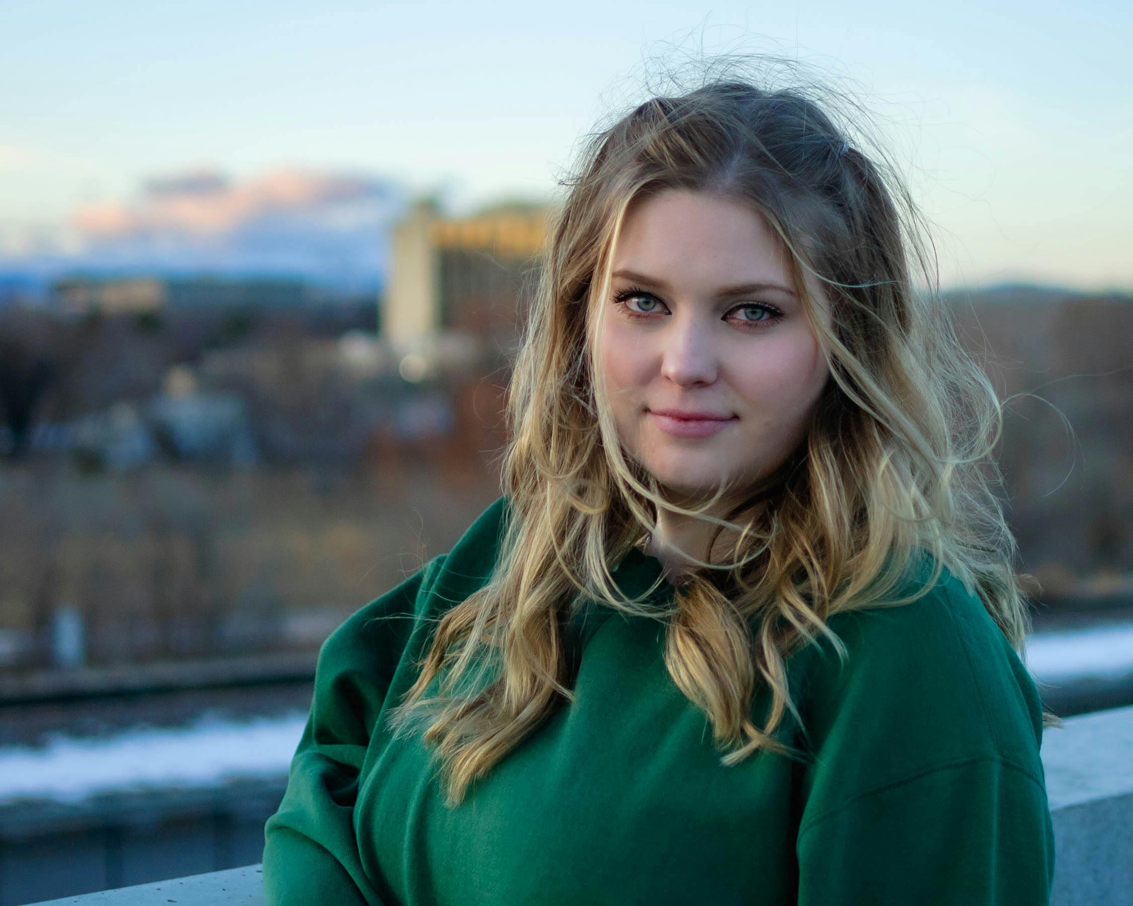 Blonde woman in a green sweater with wavy hair posing outdoors during the day.