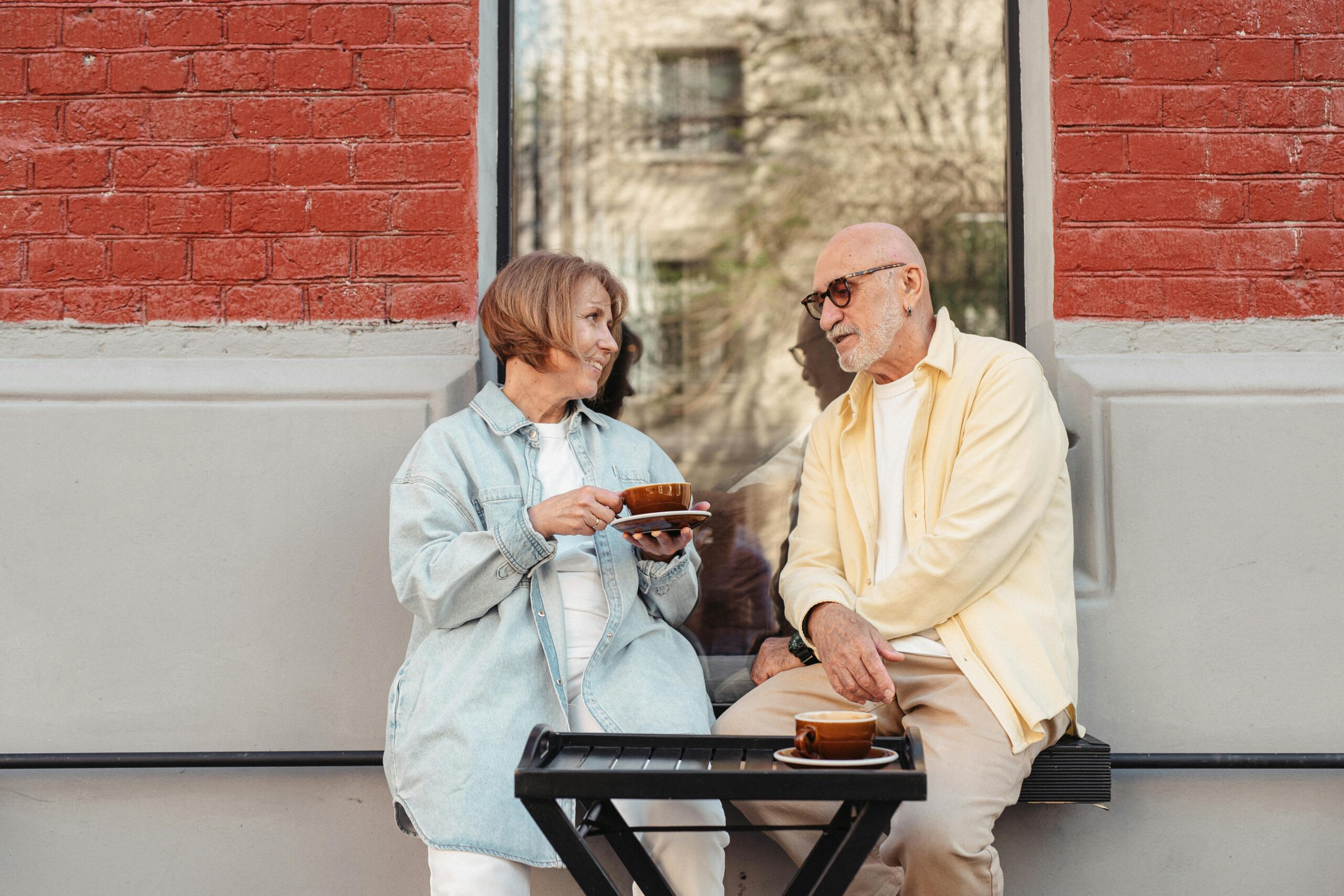 Home Senior couple sitting outdoors, enjoying coffee against a brick wall.
